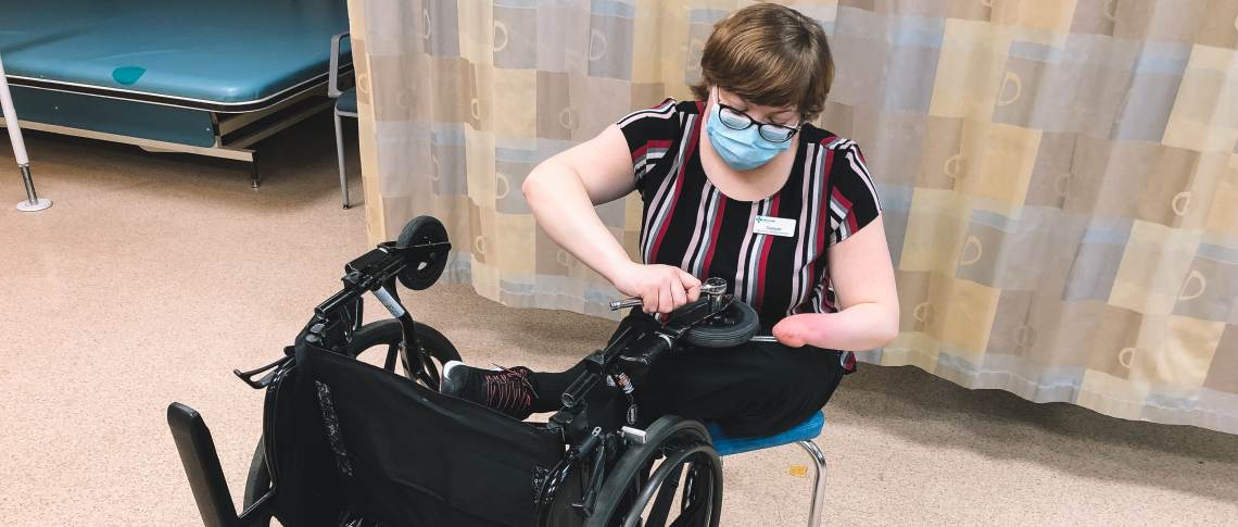 Sawyer, a white woman with a left-hand amputation, short hair, a blue mask, and glasses sits in front of a tipped over wheelchair. She is repairing the caster wheel on the wheelchair using a wrench and ratchet.