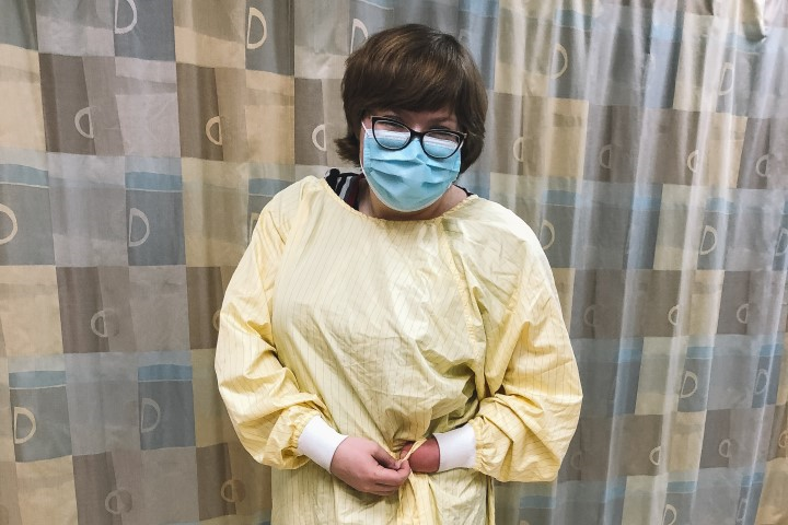 Sawyer, a white woman with a left-hand amputation, short hair, a blue mask, and glasses ties up a yellow isolation gown while smiling toward the camera.