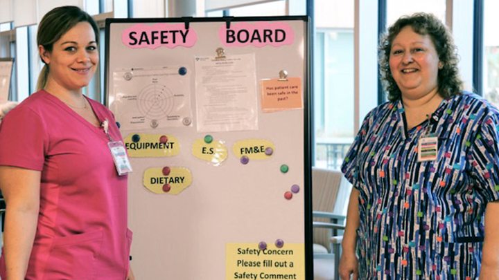 Health Care Aides Nicole Woods, left, and JoAnne Derouard pose by a patient safety board that shares safety news and invites comments from frontline staff on how to further improve safety at Edson Continuing Care Centre.